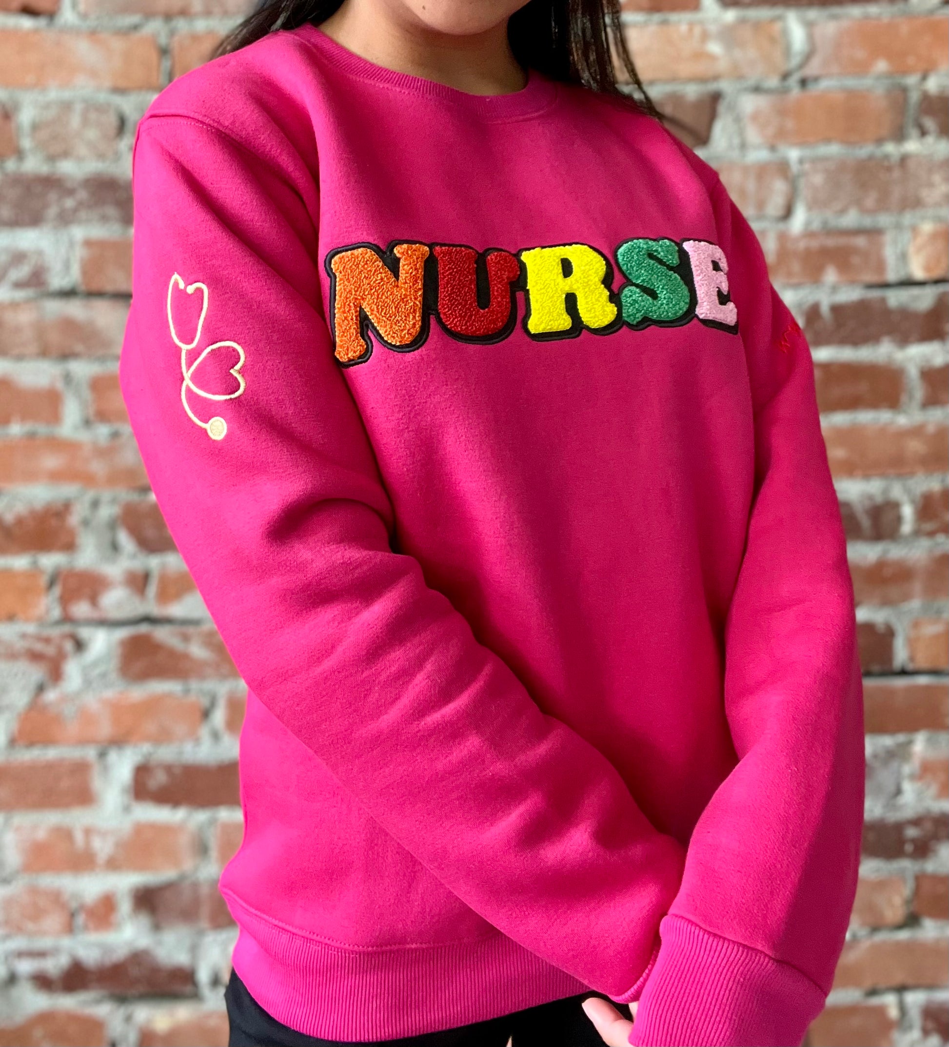 A person wearing a pink sweatshirt with the word 'NURSE' embroidered in colorful letters on the front.